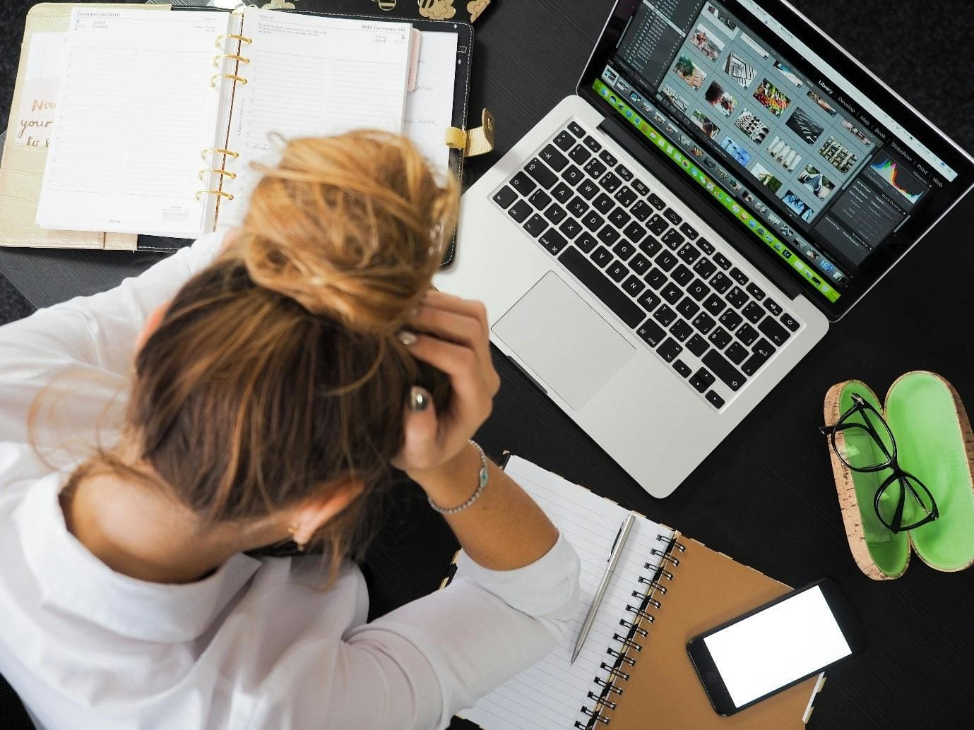 A woman sits at a desk with a laptop, notebook, organiser and glasses. She has her head in her hand looking stressed.