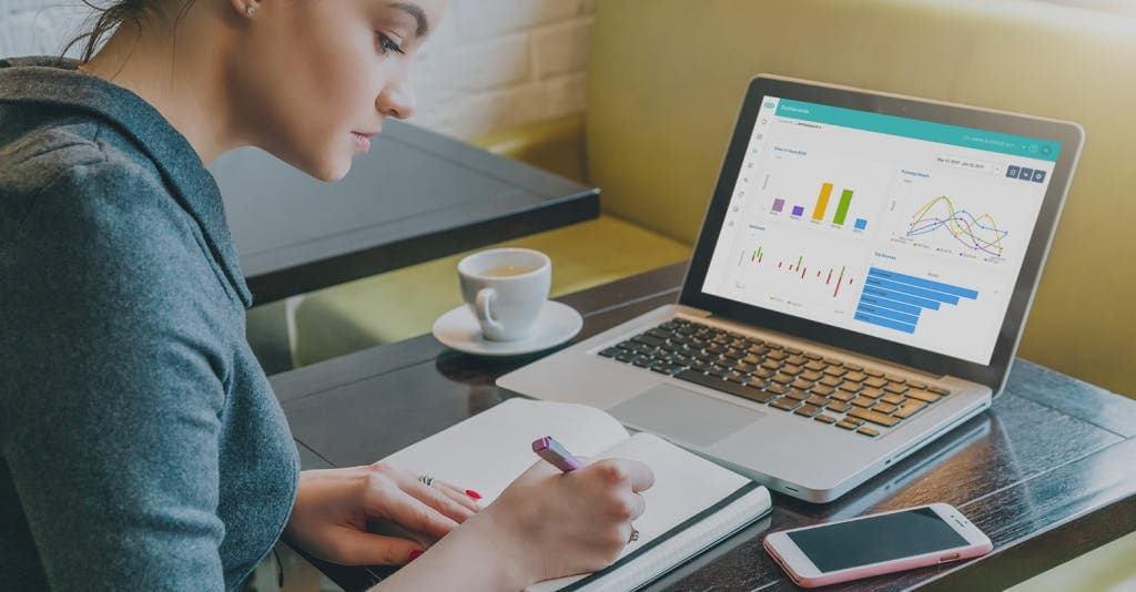 woman writing in notebook with open laptop, coffee cup and iphone on desk