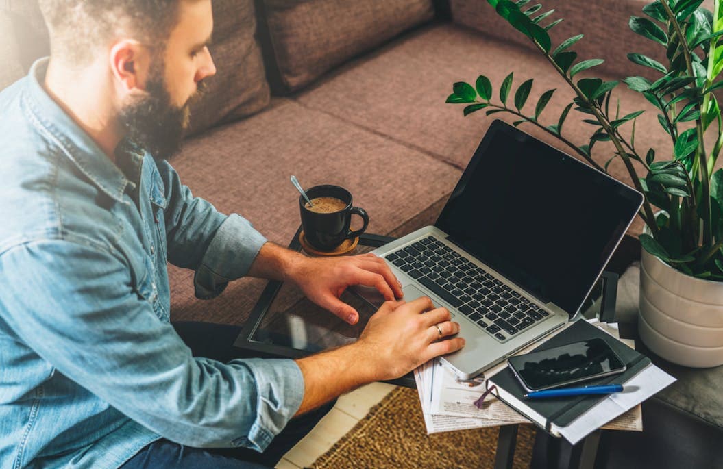 A man sits at his desk working on his laptop. On top of the desk there's also a cup of coffee, notebooks and pens, as well as a plant.