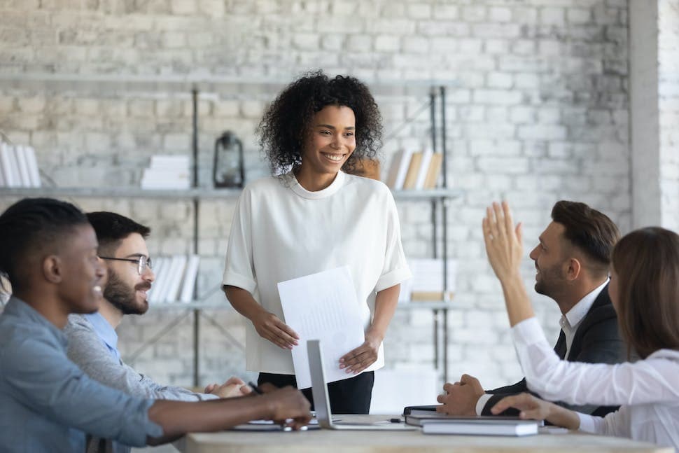 A woman holding some paper, standing infront of a group of people while one raises her hand.