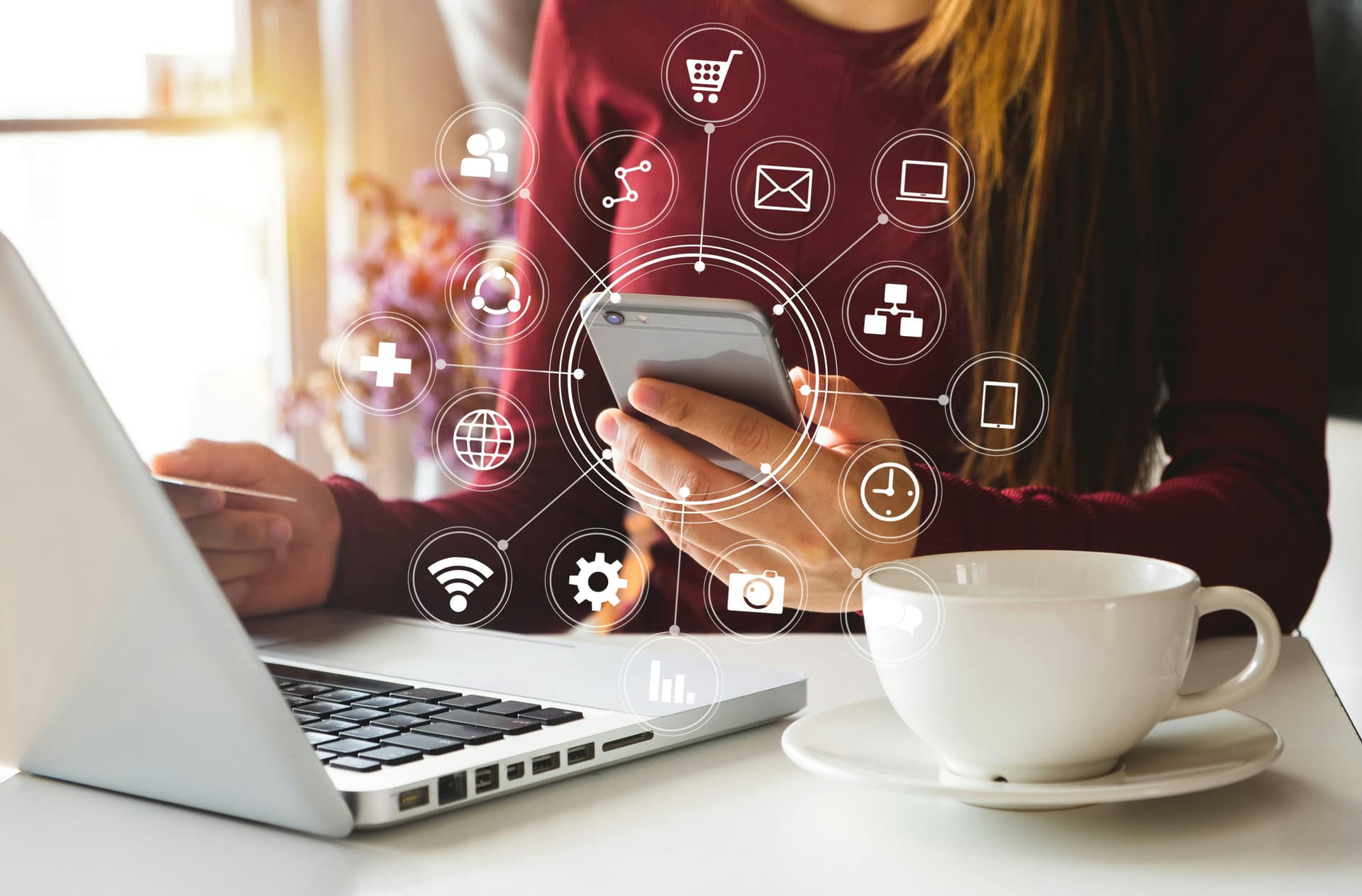 Person sitting at a laptop with cup of coffee and a phone displaying a variety of digital apps