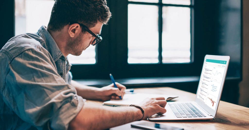 A man working on a laptop featuring graphs, whill he writes on paper
