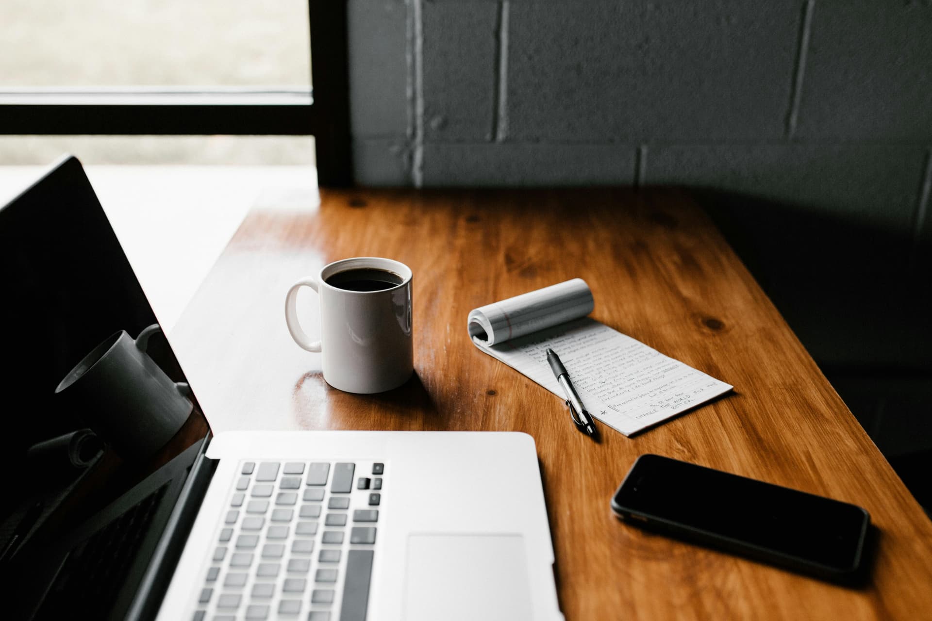 laptop on wooden desk with coffee mug, phone, and writing pad
