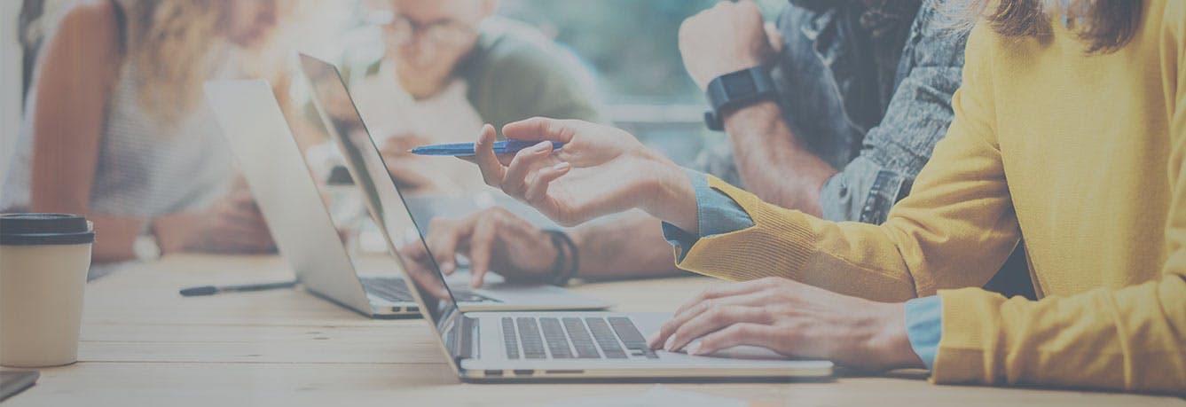 People sitting at a table working. A woman in a yellow jumper points at a laptop while a man sits next to her looking on.